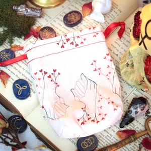 May include: A white fabric drawstring pouch featuring a black line art illustration of two hands reaching toward each other, accented with small red heart-shaped flowers on thin stems. The pouch has a red ribbon closure and a repeating red heart pattern.
