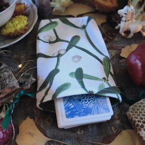 Könnte beinhalten: Eine weiße Tarotkarten-Tasche mit grünem Mistel-Muster, die ein Kartenspiel enthält. Die Tasche ist von dekorativen Gegenständen umgeben, darunter ein Granatapfel, eine Kerze und eine kleine Einhornfigur.