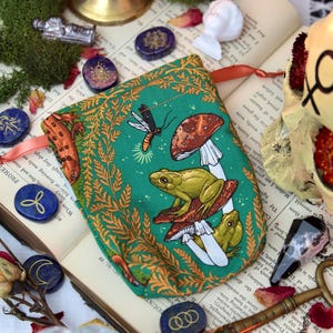 May include: A green pouch with a frog and mushroom design, accented with orange and white details. The pouch is surrounded by open books, stones, and dried roses. A skull and key are also visible.