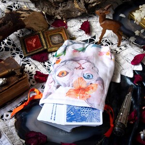 May include: A white fabric drawstring pouch featuring a watercolor illustration of a cat with one orange eye and one blue eye, adorned with orange flowers. The pouch contains a deck of playing cards with blue and white patterned backs.