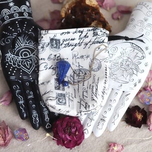 May include: A black and white decorative hand holds a small pouch with a blue gemstone and a silver chain. The pouch is printed with postage stamps and script. Rose petals and fairy lights surround the hands.
