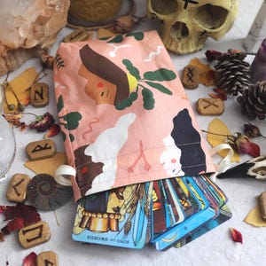 May include: A peach-colored fabric drawstring pouch featuring a stylized graphic print of faces and abstract shapes, containing a deck of tarot cards with the King of Swords visible, surrounded by candles, crystals, runes, and a bird skull.