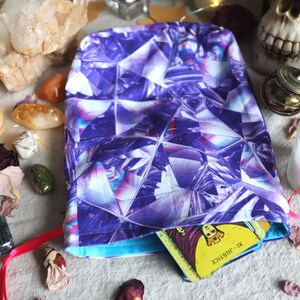 Op de afbeelding: Een paarse trekkoordtas met een geometrisch diamantpatroon, met een tarotkaart. De tas is omgeven door kristallen, kaarsen en gedroogde rozenblaadjes. Een schedel en andere spirituele items staan op de achtergrond.