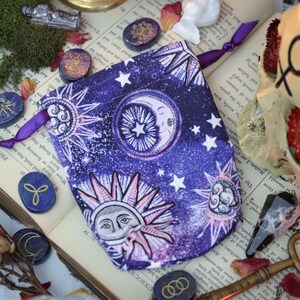 May include: A purple drawstring bag with a celestial design featuring suns, moons, and stars. The bag is surrounded by blue and gold rune stones, dried roses, and a skull on an open book. The bag has a purple ribbon closure.