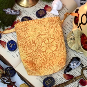 May include: An open book with a yellow floral patterned pouch, blue stones, dried roses, and a decorative skull. The pouch has a drawstring closure. The stones have gold designs. The skull has black markings.