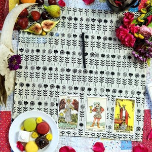 May include: An overhead shot features a goat skull adorned with flowers, a floral-patterned cloth, and a deck of tarot cards. The scene includes fresh fruit, macarons, and rose petals scattered on a colorful quilt, creating a mystical aesthetic.
