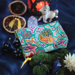 May include: A teal dice bag with a colorful floral and abstract pattern, open to reveal dice. The scene includes a candle, a unicorn figurine, dried orange slices, and a blue crystal pendant, all on a blue fabric surface.