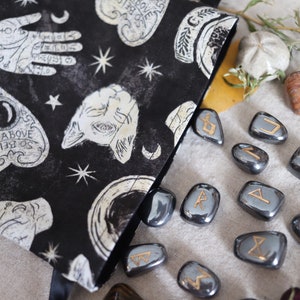 May include: Black fabric bag with white celestial symbols and the text "AS ABOVE SO BELOW". A collection of gray rune stones with gold symbols are scattered around the bag. Decorative stones and dried flowers are also present.