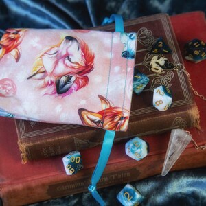 Pode incluir: Uma bolsa de dados rosa com uma ilustração de raposa, cheia de dados variados, repousa sobre livros antigos. Um pêndulo de cristal transparente e uma fita azul adicionam elementos decorativos. Fundo de veludo azul escuro.