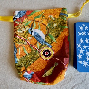 May include: A fabric tarot card bag with a colorful bird and floral design in shades of orange, red, and yellow. A stack of blue tarot cards with white star patterns is next to the bag. A yellow ribbon is tied to the top.