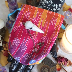 May include: A vibrant pouch with a crystal pendulum, a bird skull, and a lit candle. The pouch features a hand-drawn pattern in pink, orange, and purple. The pendulum has a silver chain and a small pink bead. Various crystals and dried flowers are also present.