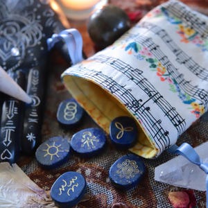 May include: An assortment of mystical items. Blue rune stones with gold symbols are scattered near a pouch decorated with musical notes and flowers. A crystal and a black hand with white markings are also present. The items are on a patterned surface.