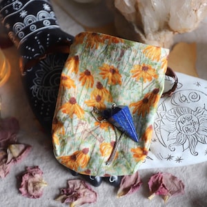 May include: A blue gemstone pendulum with a silver chain rests on a floral print pouch. The pouch features orange and yellow flowers on a green background. Decorative candles and a black and white painted object are also in the image.