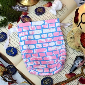 May include: A pink and white brick-patterned drawstring bag with a white ribbon, surrounded by blue and gold stones, dried roses, and a skull. The bag is on an open book with text.