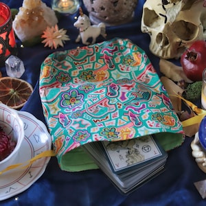 May include: A teal tarot card bag with a colorful floral and geometric pattern, filled with a deck of cards. The scene includes candles, crystals, dried fruit, and a skull on a blue cloth, creating a mystical aesthetic.