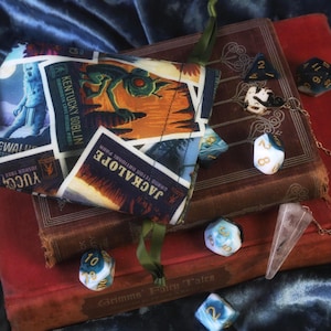 May include: A dice bag with travel-themed graphics, placed on old books. The bag displays images of a goblin and the text "Kentucky Goblin" and "Jackalope". Several dice, a crystal pendulum, and a chain are arranged around the books.