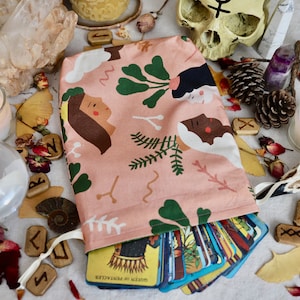 Puede incluir: Una bolsa rosa para cartas del tarot con un patrón de caras y follaje ilustrados. La bolsa está abierta, revelando una baraja de cartas del tarot. La imagen también incluye cristales, velas, runas y una calavera, lo que sugiere un tema espiritual o místico.