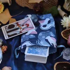 May include: A navy blue tarot card bag with a celestial pattern, holding a deck of cards. A tarot card with swords is placed next to the bag. The scene includes dried flowers, crystals, and a pomegranate on a dark blue surface.