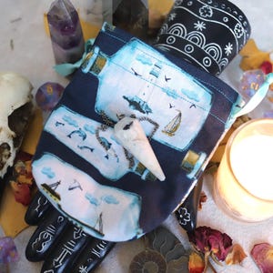 May include: A dark blue drawstring pouch with a nautical design featuring sailboats and bottles. A white pendant hangs from the pouch. The image also includes a bird skull, crystals, dice, a candle, and a black and white patterned container.