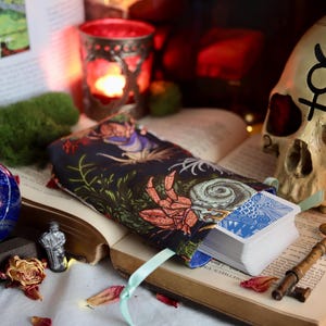 Puede incluir: Una naturaleza muerta mística con una baraja de cartas del tarot en una bolsa con estampado floral, una calavera, una esfera azul y una llave vintage. Los objetos están dispuestos sobre un libro abierto con flores secas y una vela encendida.