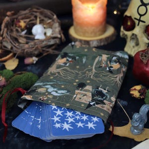 May include: A green tarot card bag with a whimsical forest design, holding a deck of blue cards with white star patterns. The scene includes candles, a skull, and a crystal ball, creating a mystical ambiance.