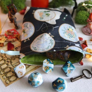 Puede incluir: Una bolsa de dados negra con diseños florales bordados, llena de dados. Varios dados poliédricos en tonos azules y blancos están esparcidos alrededor de la bolsa, junto con figuras en miniatura y una llave. Una vela encendida y pétalos secos completan la escena.