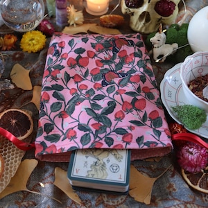 Pode incluir: Uma bolsa rosa para cartas de tarot com um padrão de morangos e folhas, contendo um baralho de cartas. A imagem inclui uma xícara de chá branca com um cristal, uma caveira, velas e frutas secas, criando uma estética mística.
