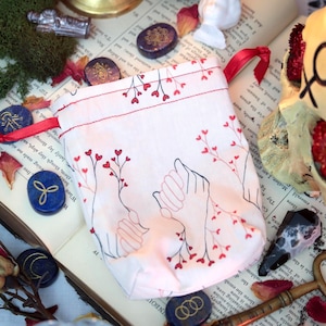 May include: A white drawstring pouch with a red heart and hand-holding design. The pouch is on an open book with other decorative items, including stones, dried flowers, and a skull.
