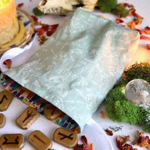 Pode incluir: Uma bolsa de tecido azul claro com um padrão floral branco, revelando um interior colorido. Pedras rúnicas de madeira e figuras de pássaros pretos estão espalhadas ao redor da bolsa. Uma vela acesa e uma bola de cristal também estão presentes.