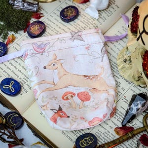 May include: A drawstring pouch with a woodland scene featuring a deer, birds, and mushrooms. The pouch is cream-colored with a pink ribbon. The image also includes decorative stones, dried roses, and a skull.