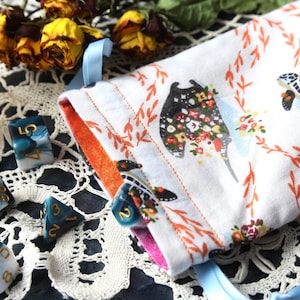 May include: A white fabric drawstring pouch featuring a repeating pattern of manta rays and sea turtles decorated with floral motifs, accompanied by several polyhedral dice with a blue and white marbled finish and gold-colored numbers.