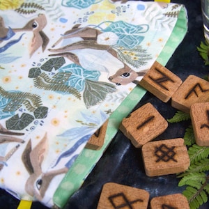 May include: A patterned fabric featuring deer and floral designs, with wooden rune stones bearing etched symbols. The fabric has a green border. A glass of water and fern leaves are also visible.