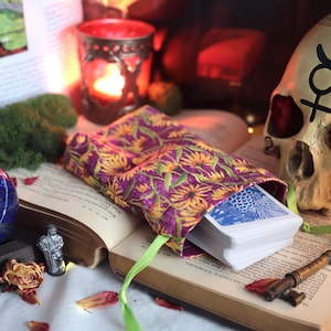 May include: A purple and yellow floral tarot card bag with a deck of cards peeking out, resting on an open book. A skull, a blue sphere, and a key are also present, suggesting a mystical or occult theme.