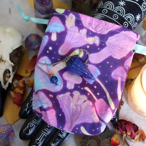 May include: A purple drawstring bag with a mushroom pattern, holding a blue crystal pendulum. The scene includes a bird skull, dice, and a lit candle, creating a mystical aesthetic. The bag is tied with a light blue ribbon.