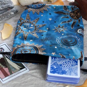 Könnte beinhalten: Eine blaue Tarotkarten-Tasche mit einem Sonnen- und Sternenmuster. Ein Stapel blau-rückseitiger Karten ist teilweise sichtbar. Eine Tarotkarte mit Illustration liegt neben der Tasche. Eine brennende Kerze und Kristalle im Hintergrund.