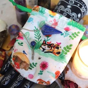 May include: A white drawstring bag with a colourful floral and cat print, holding a blue crystal pendulum and a silver chain with a blue bead. A skull, candle, and other items are in the background.