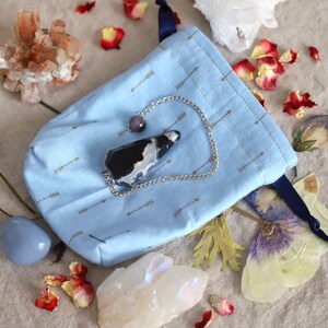 May include: A light blue pouch with a silver chain and a black and white crystal pendant. The pouch has a pattern of small arrows. Dried flowers and crystals are scattered around the pouch.