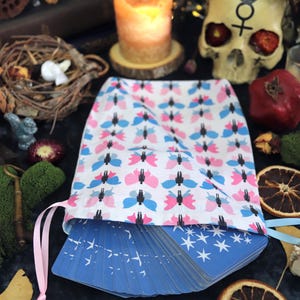 May include: A white drawstring bag with a pink and blue butterfly pattern, filled with blue tarot cards. The cards have white star designs. The bag is surrounded by candles, a crystal ball, and other mystical items.