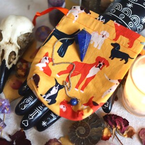 May include: A yellow drawstring bag with a dog pattern holds a blue crystal pendulum. A raven skull and a lit candle are also in the image, along with other crystals and dried flowers. The theme is spiritual and mystical.
