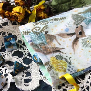 May include: A fabric drawstring pouch featuring a printed pattern of deer, ferns, and yellow flowers, paired with a set of multi-faceted polyhedral dice in shades of teal, white, and blue with gold-coloured numbers, arranged on a white lace surface.