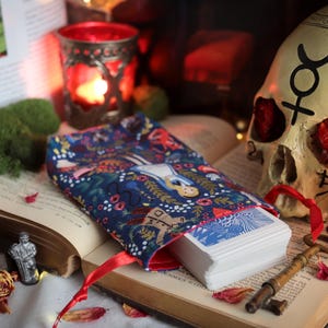 May include: A deck of cards in a blue floral patterned pouch with a red ribbon, resting on an open book. A skull with markings and a candle holder are in the background. A blue sphere and a small figurine are also visible.
