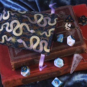 Op de afbeelding: Een donkerblauwe dobbelsteenzak met een slangenpatroon, rustend op antieke boeken. Verschillende dobbelstenen in blauw, wit en zwart liggen verspreid rond de boeken. Een heldere kristallen pendel hangt in de buurt. De scène speelt zich af op een donkerblauwe fluwelen ondergrond.
