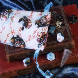 May include: A white dice bag with a floral and turtle pattern, accented with a light blue ribbon, rests on antique books. Various polyhedral dice in different colors and a crystal pendulum are scattered around the books, suggesting a tabletop gaming theme.