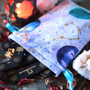 May include: A white drawstring bag with a celestial design, holding black wooden runes with gold symbols. The bag is decorated with blue and gold constellations. A lit red candle in a decorative holder and dried rose petals are in the background.