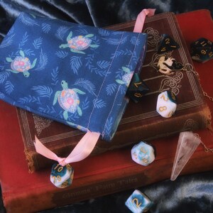 May include: A blue fabric dice bag with a turtle pattern, resting on antique books. Several dice of various colors and a crystal pendulum are scattered around the books. A pink ribbon is tied to the bag.