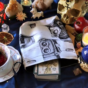 May include: A white tarot card bag with a black camera illustration, filled with tarot cards, sits on a blue surface. The scene includes candles, crystals, dried fruit, a skull, and a blue sphere.