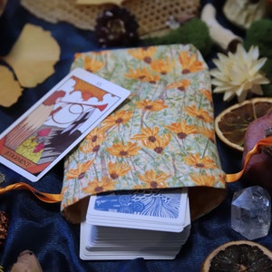 May include: A tarot card bag with a floral pattern of orange and yellow flowers on a light green background. A tarot card with the word "JUDGEMENT" is next to the bag. The bag is open, revealing a stack of tarot cards.