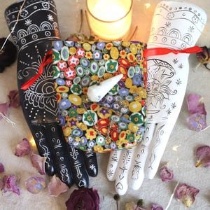 Puede incluir: Una bolsa de tela colorida con un objeto blanco en forma de diente se sostiene entre dos esculturas de manos decorativas. Una mano es negra con diseños blancos, la otra es blanca con diseños negros. Una vela y pétalos de rosa están en el fondo.