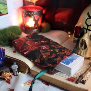 Peut inclure: Une composition de nature morte avec un jeu de cartes de tarot, une pochette à motifs floraux rouges, une sphère bleue, un crâne et une clé vintage. Les objets sont disposés sur un livre ouvert, avec une bougie en arrière-plan.