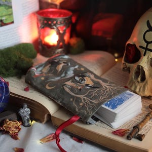 May include: An open book with a tarot card deck and a decorative skull. A blue sphere, a small figurine, and a key are also present. The tarot card deck is in a gray pouch with a forest design. A lit candle is in a red glass holder.